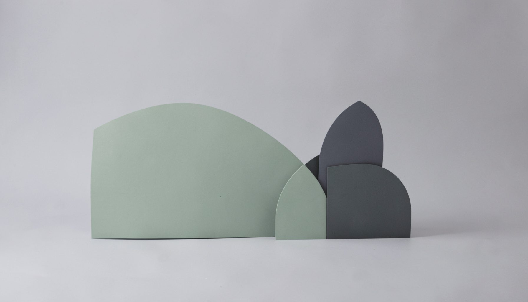 A calm studio-style photograph on a soft neutral gray backdrop, showing simple abstract geometric shapes in muted sage green and charcoal tones, evoking a quiet and orderly mood